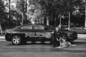 A police officer and his dog sit in front of their car.